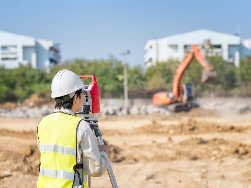 Surveyor setting layout beside a fenced school construction zone while the existing campus remains open and active.